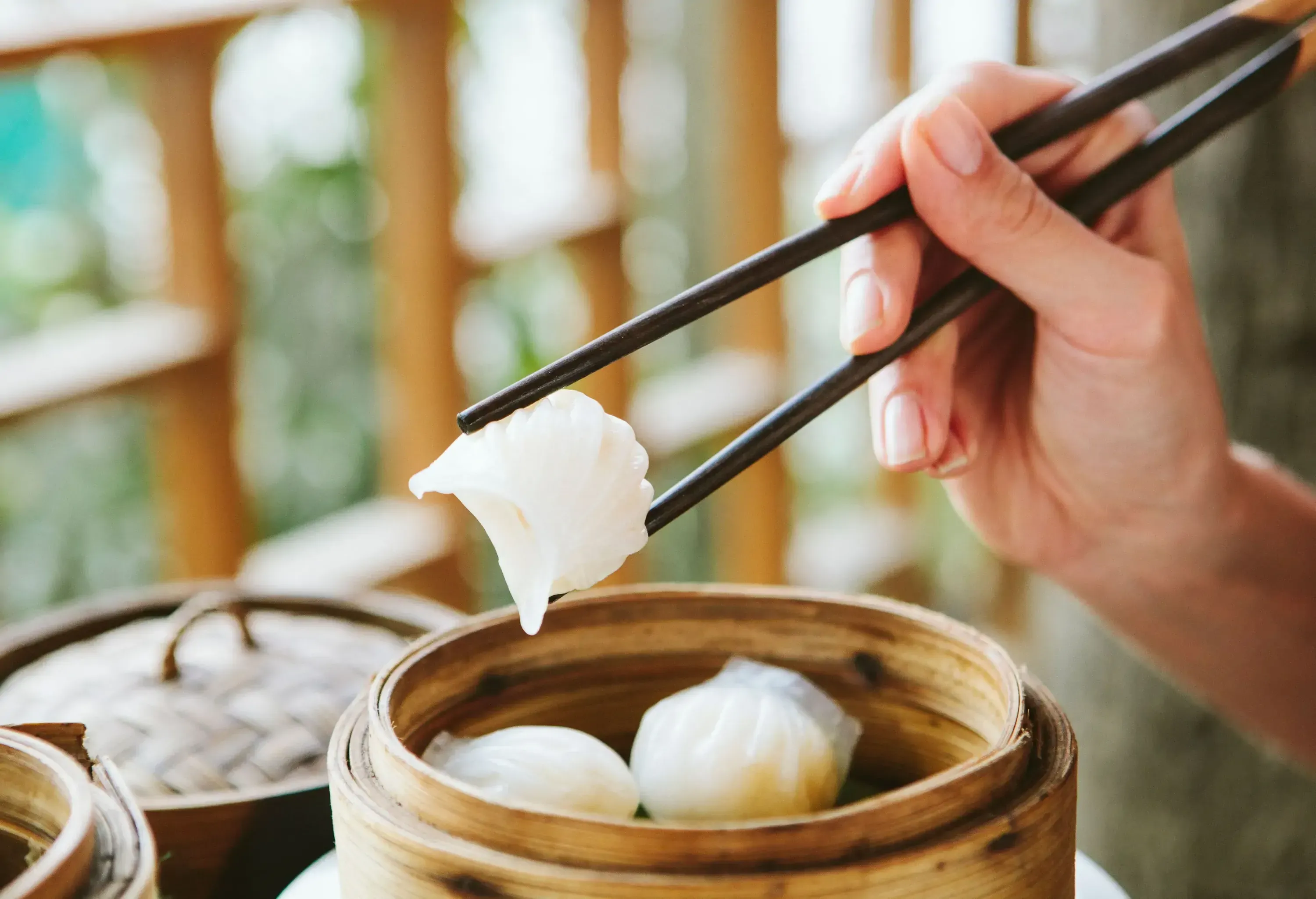 Delicious dim sum is arranged on a rustic wooden container, showcasing a hand delicately holding prawn dumplings with precision using chopsticks.