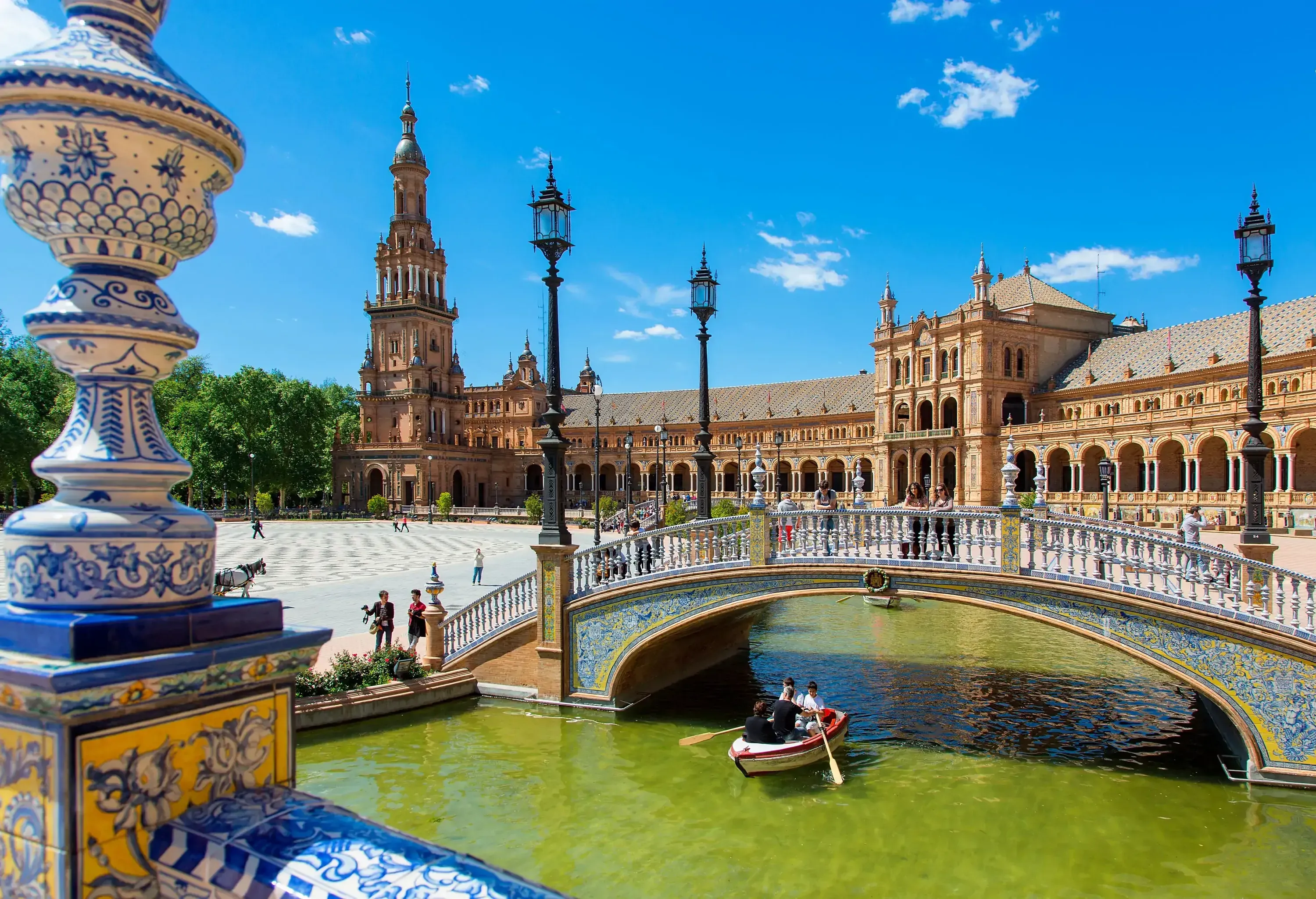 A small boat is passing beneath a gorgeously painted arched footbridge in front of Plaza de Espaa, where many people are walking.