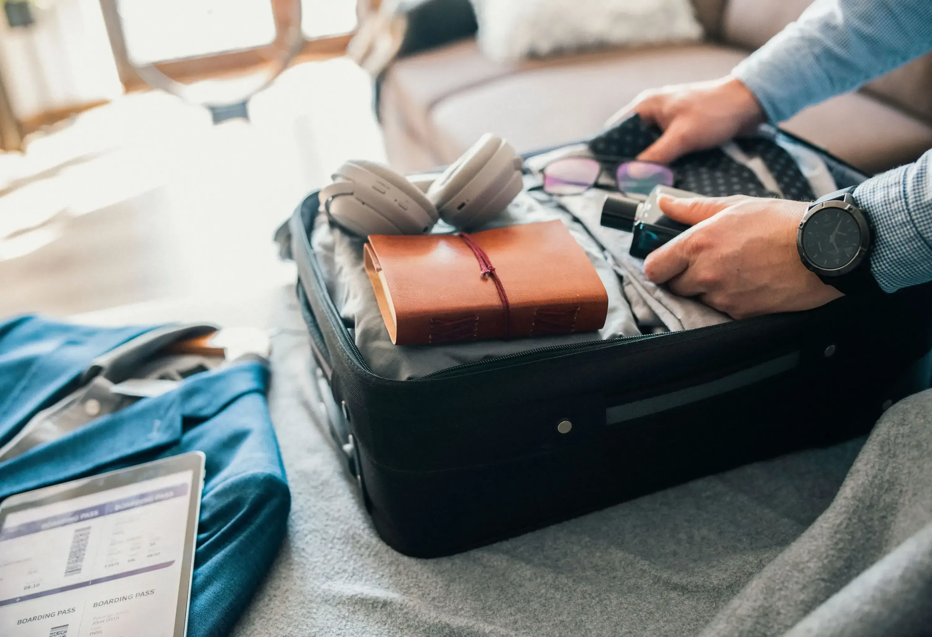 Person packing a carry-on suitcase on a bed with headphones and an orange notebook.