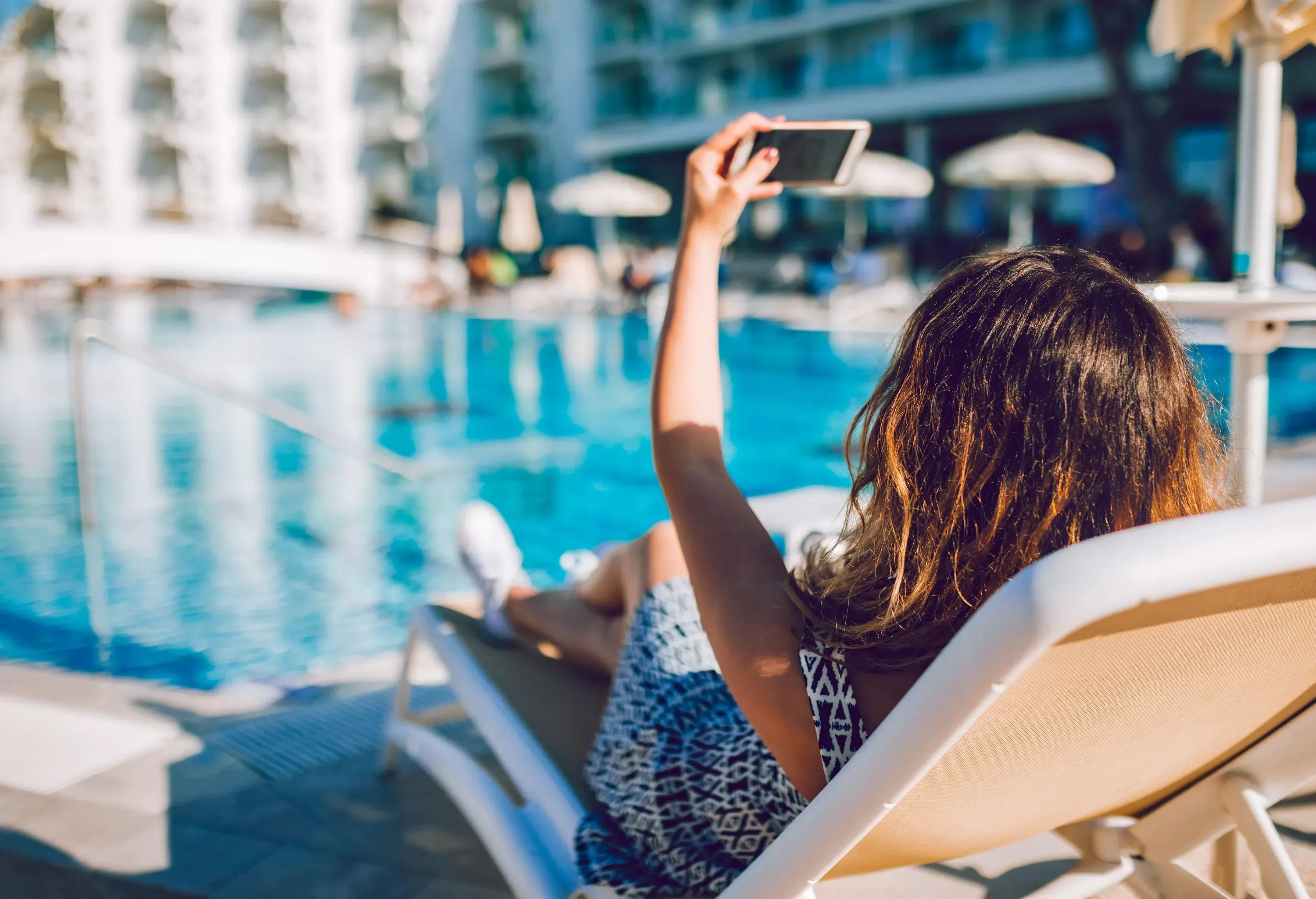 A young woman lies down on a sun lounger by a swimming pool, taking a selfie using her smartphone.