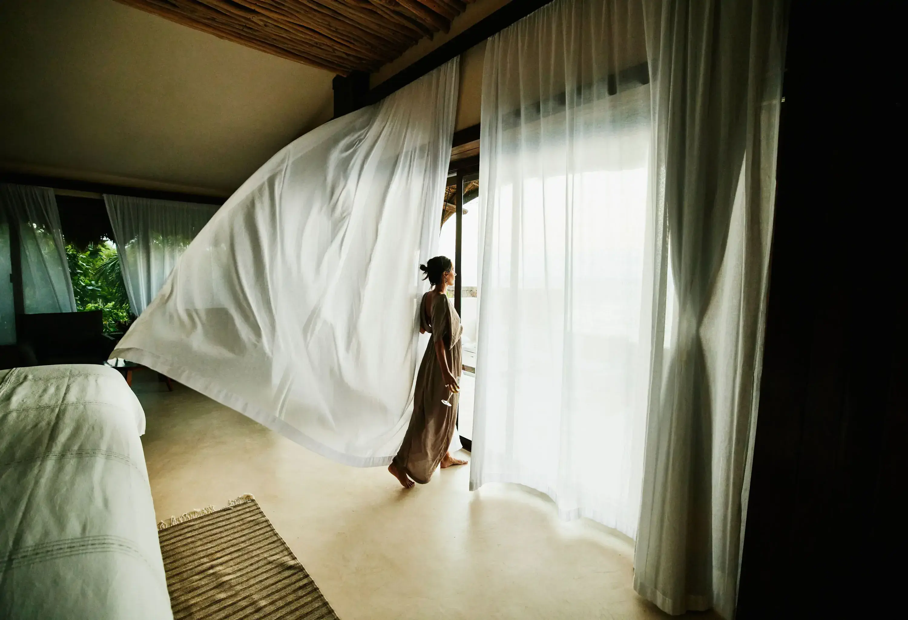 Woman in a hotel room looking out of the french windows between muslin curtains flying from the breezw