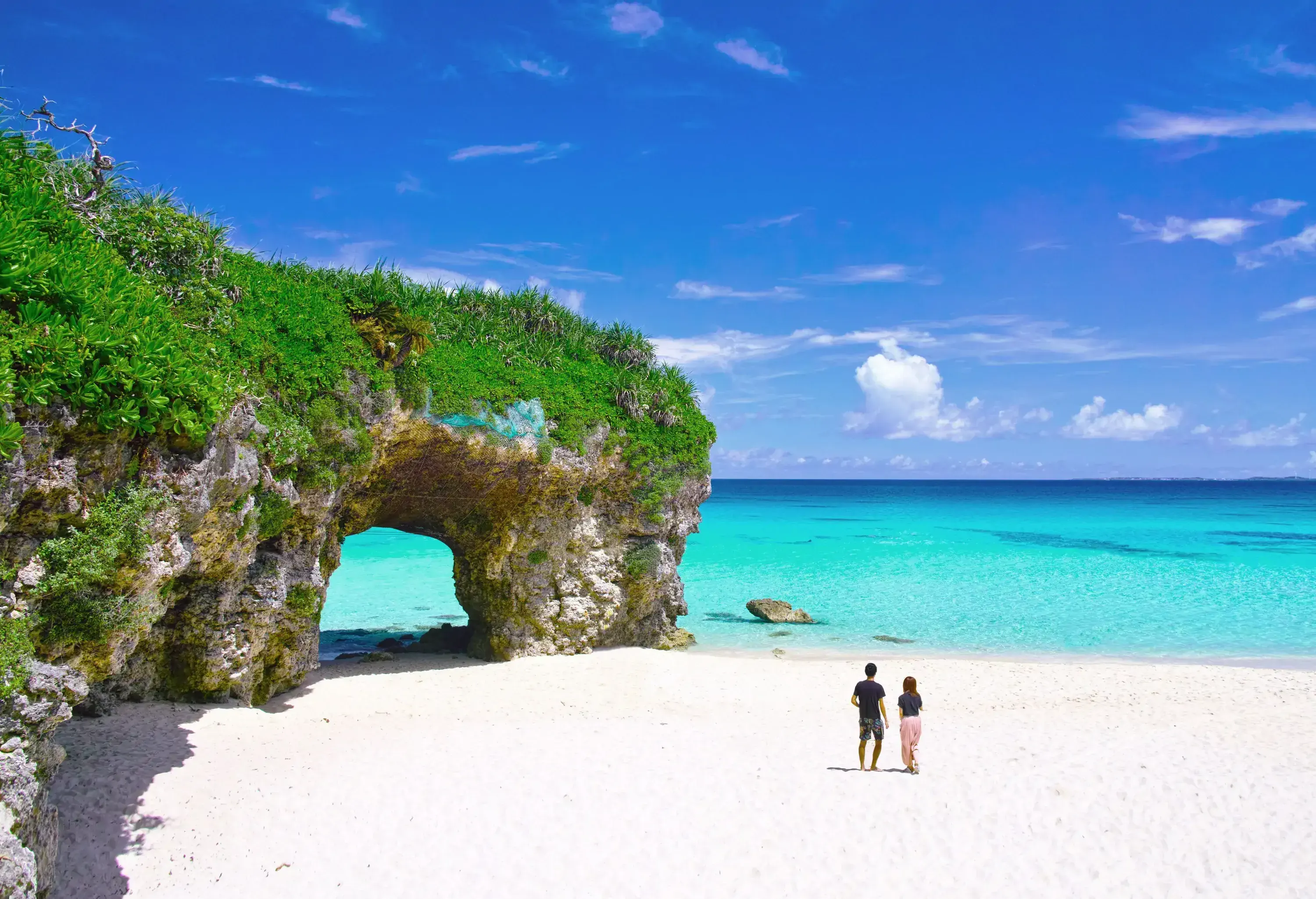 Two people walking across a sandy white beach with a natural arch.