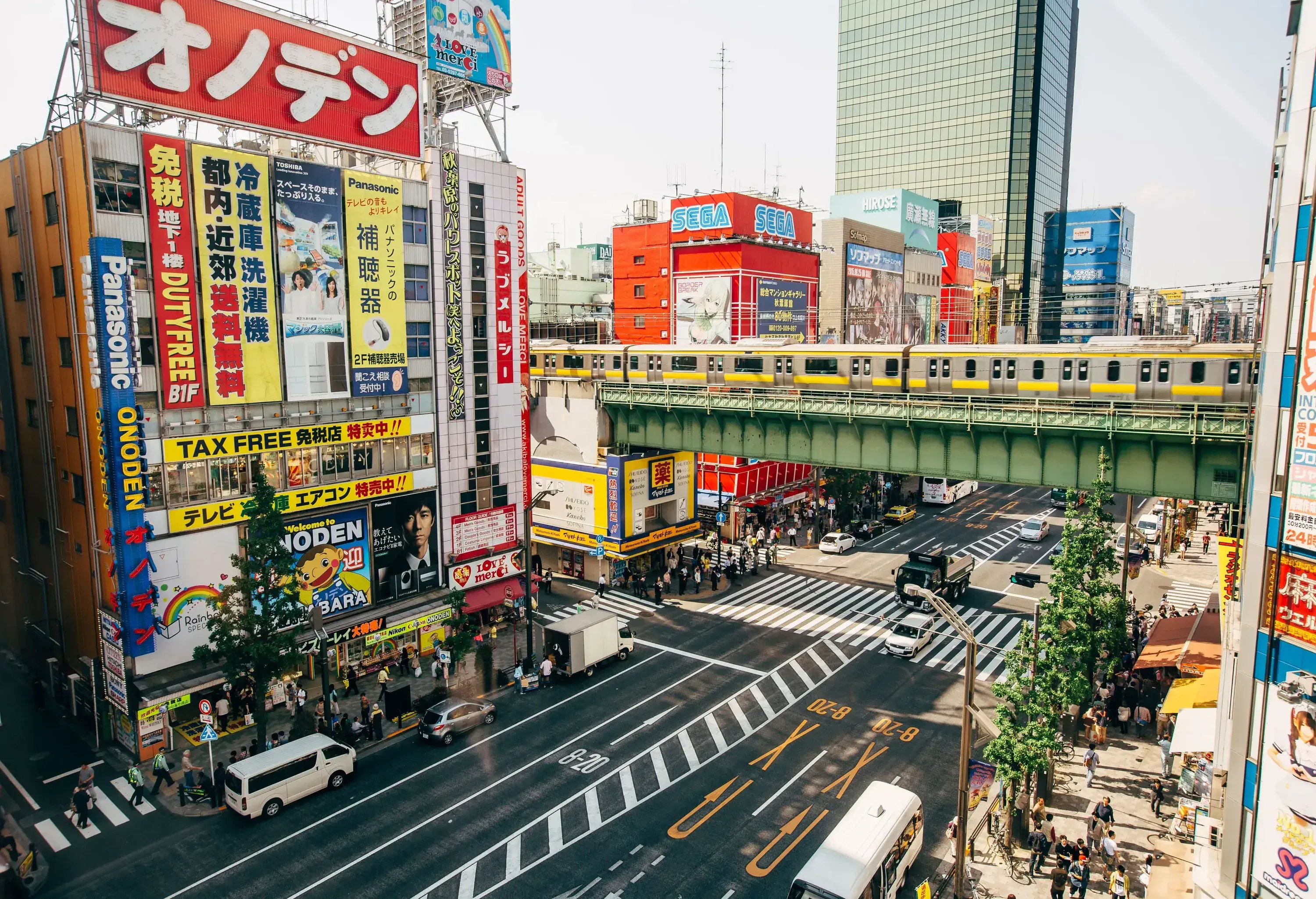 An avenue adorned with shops on both sides, featuring vibrant and colourful advertisements, while an elevated train gracefully glides along the tracks above.