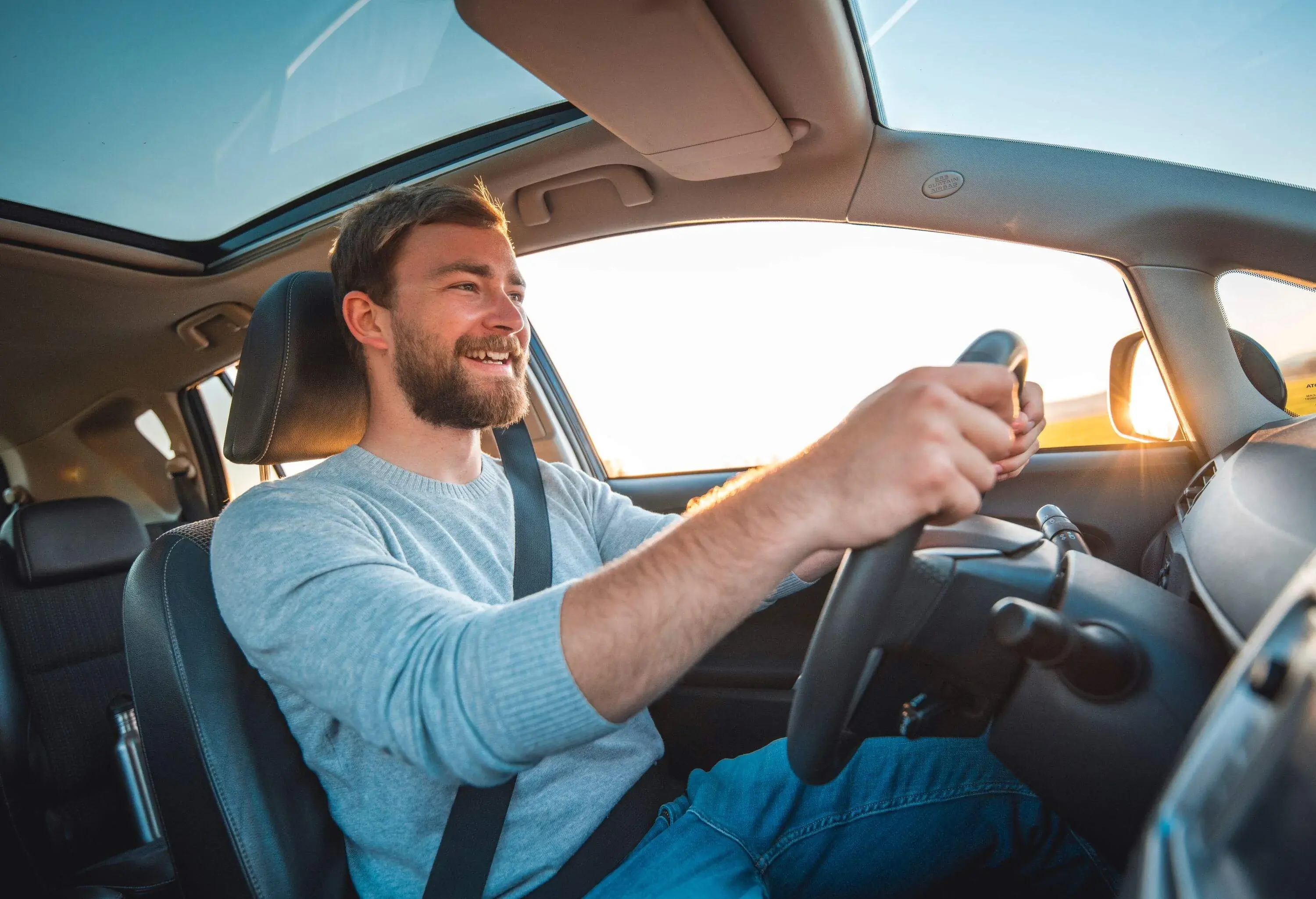 Young man in a car. He is driving, smiling and looking out. Sun is shining from back on  a sunny spring afternoon.