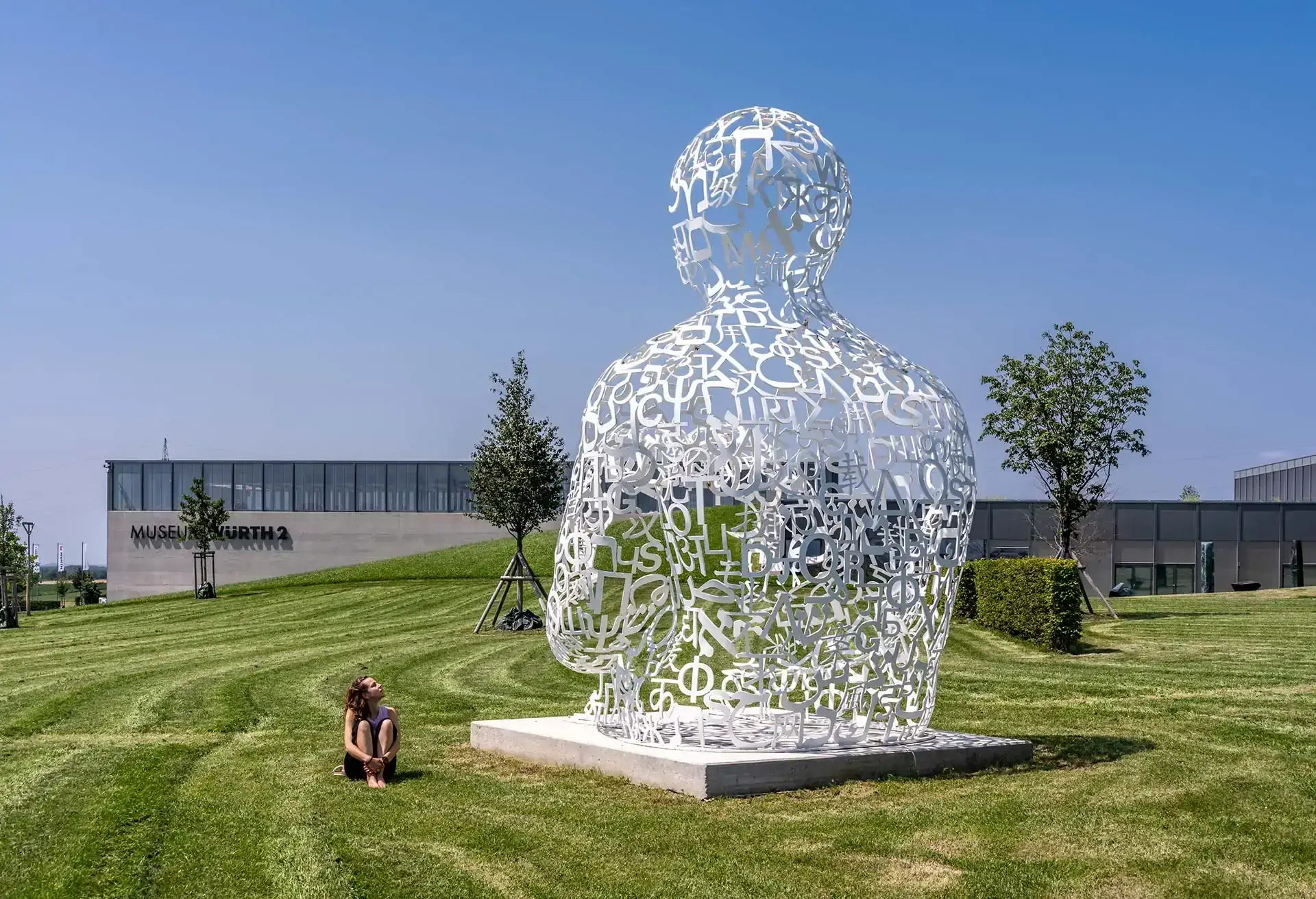 Stor hvid skulptur af en menneskefigur i en park med en person, der kigger på den.
