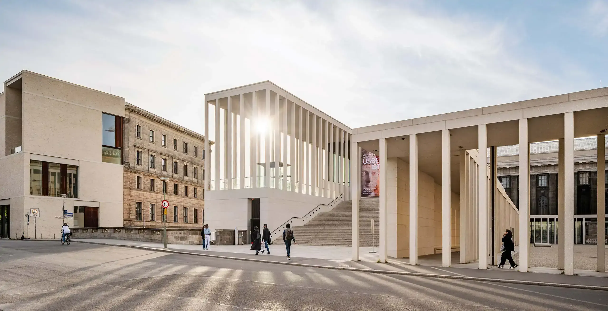 Modern and historic buildings in soft light; the sun peeks through vertical columns on the right.
