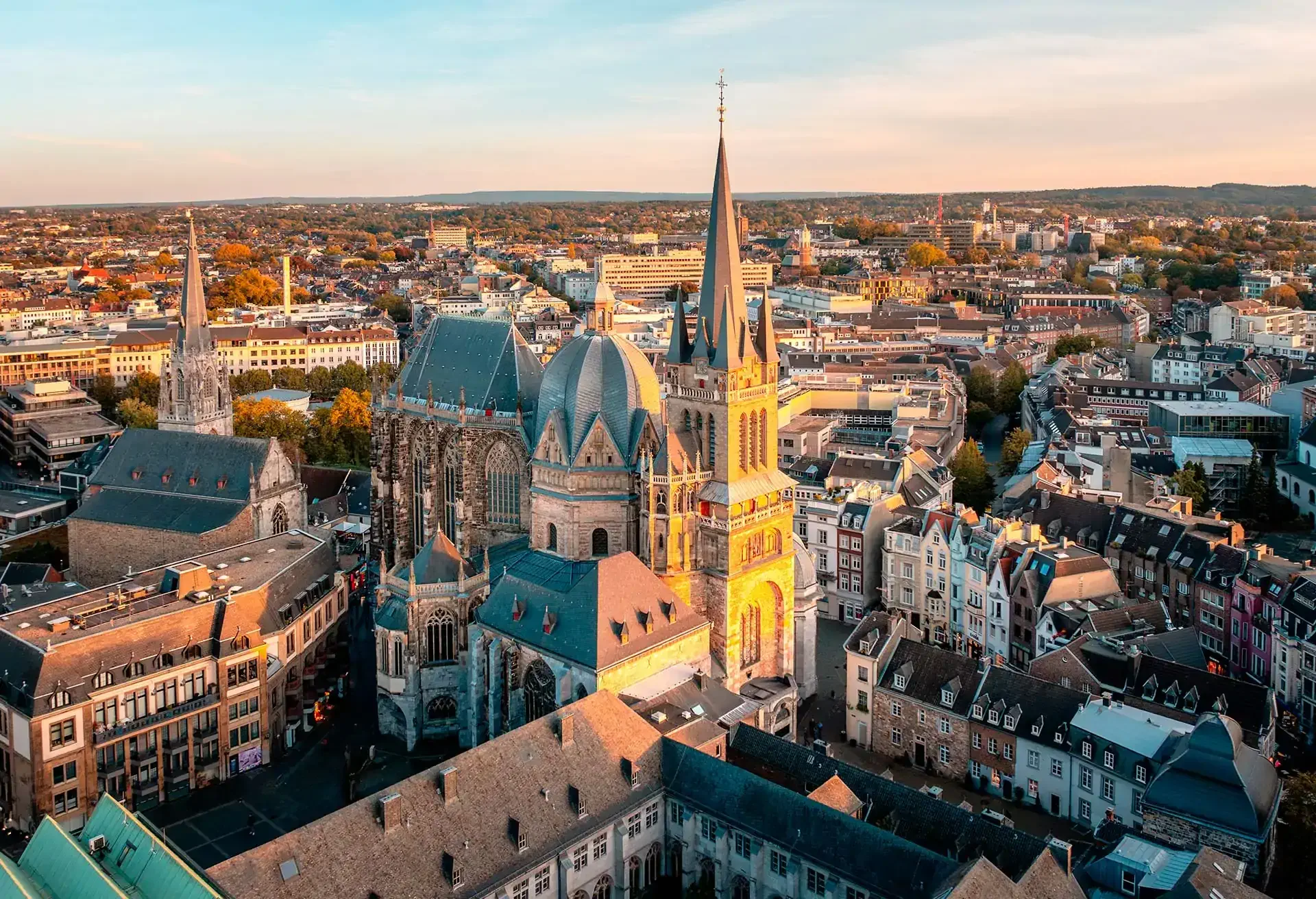 Panoramisk udsigt over Aachen med historisk arkitektur, inklusive kirke og tage ved solnedgang.