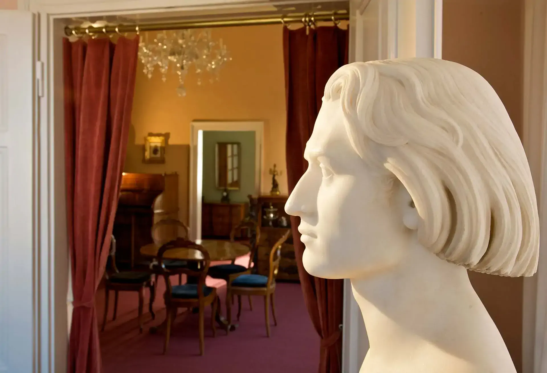 Hvid marmor bust af en mand foran en elegant værelse med møbler og lysekrone.