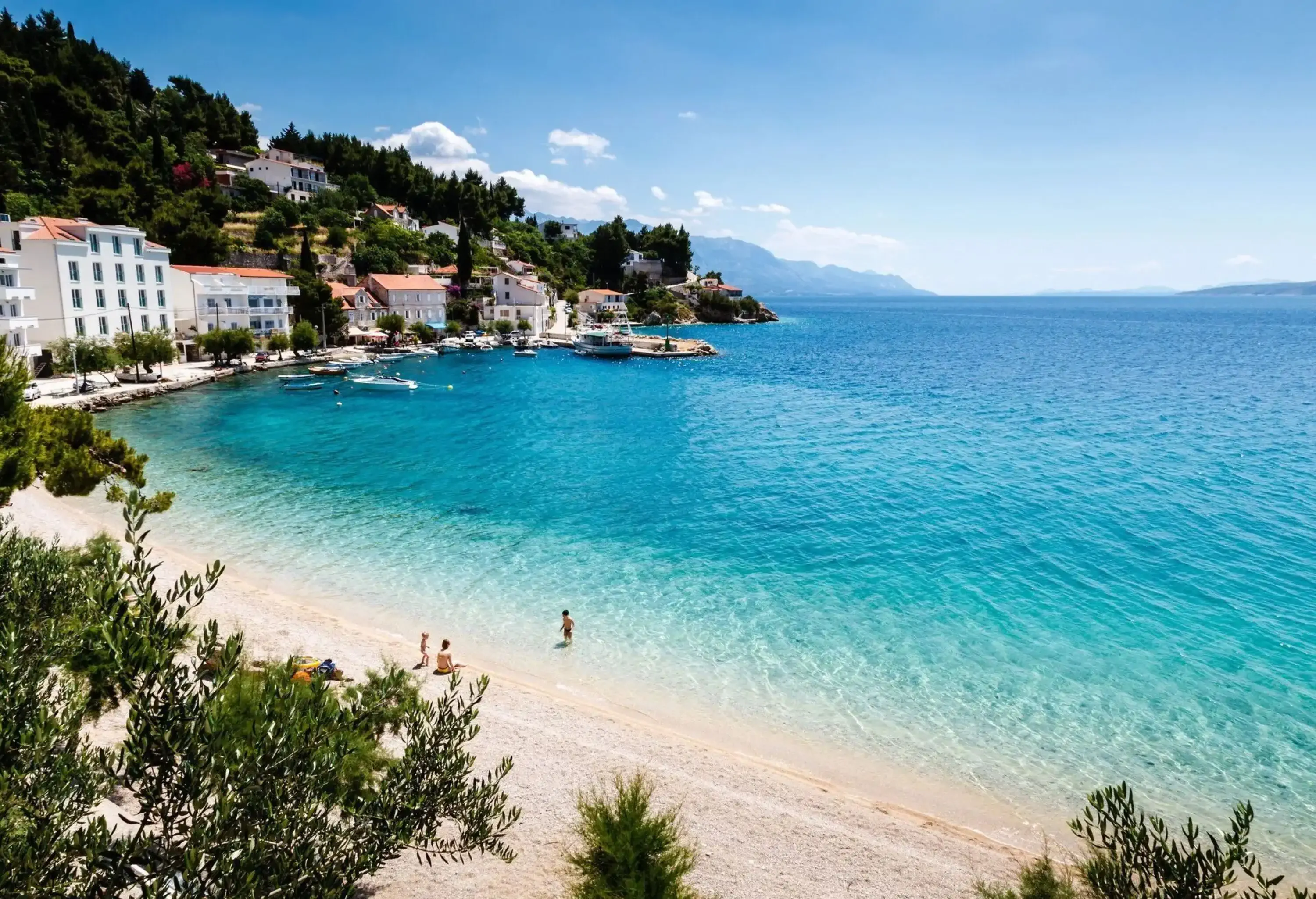 A clear water beach with white sand lined with modern buildings beneath a lush hill.