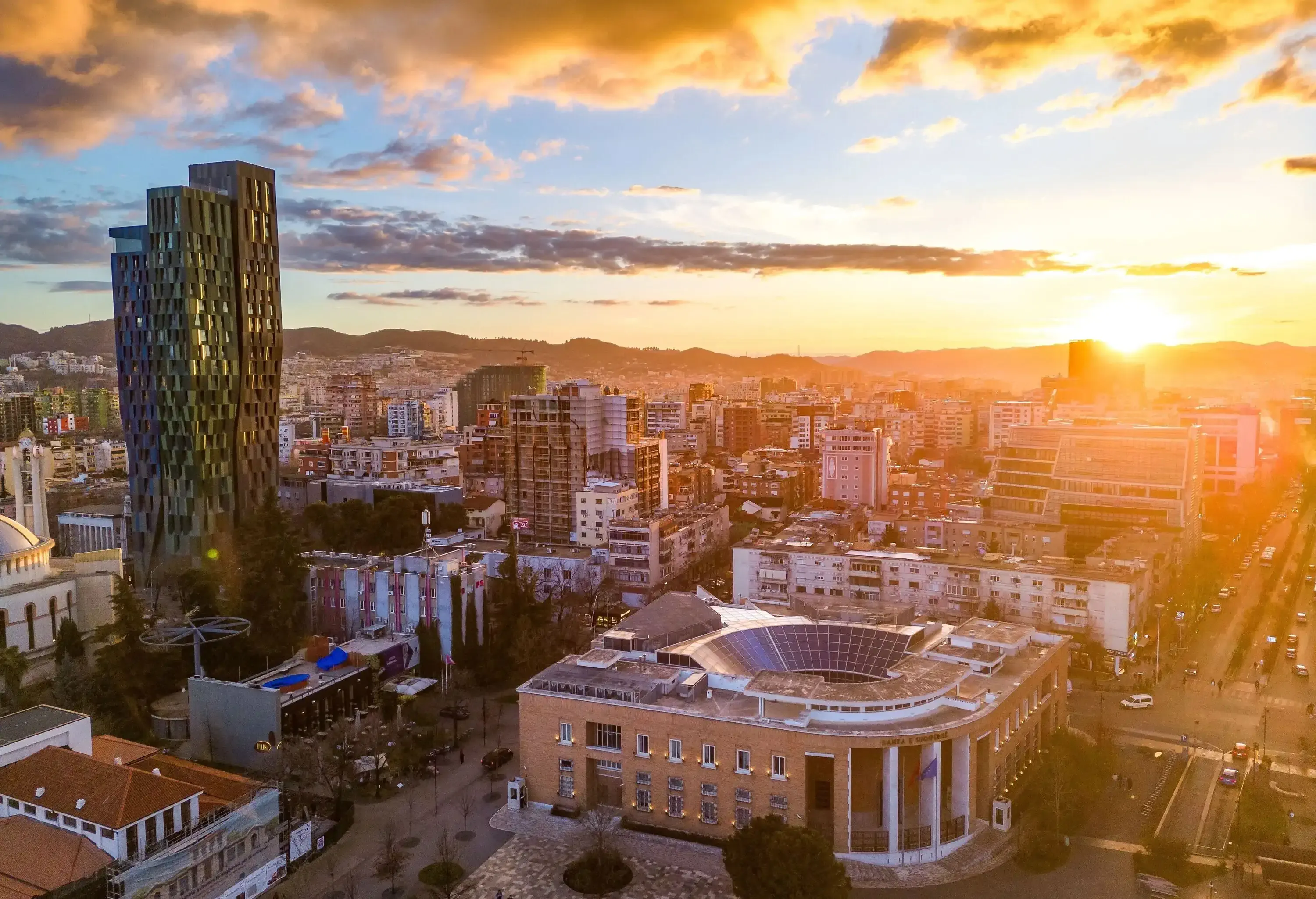 Aerial sunset view of a city blending traditional and modern architecture, backed by silhouetted mountains.