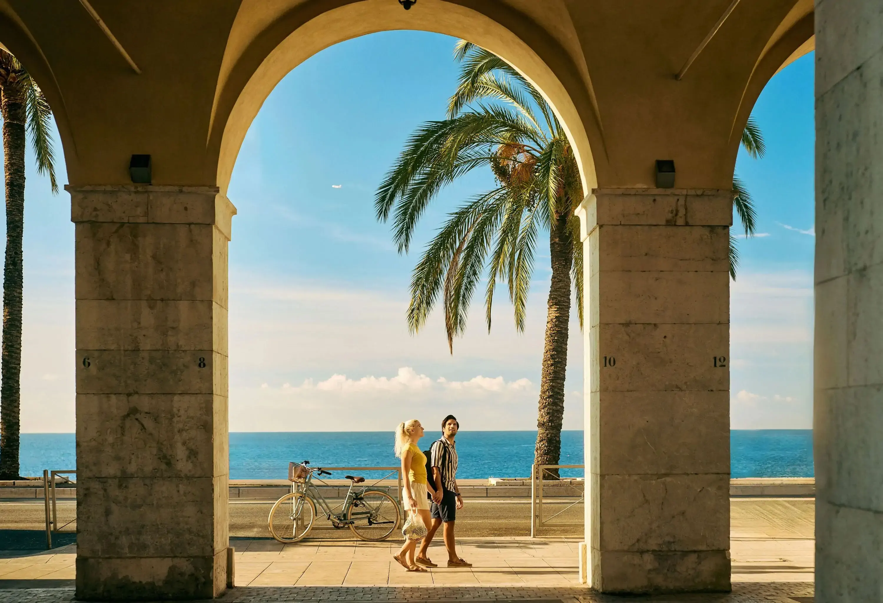 A couple walks along an oceanfront promenade framed by stone arches, with palm trees and a bicycle in the background.