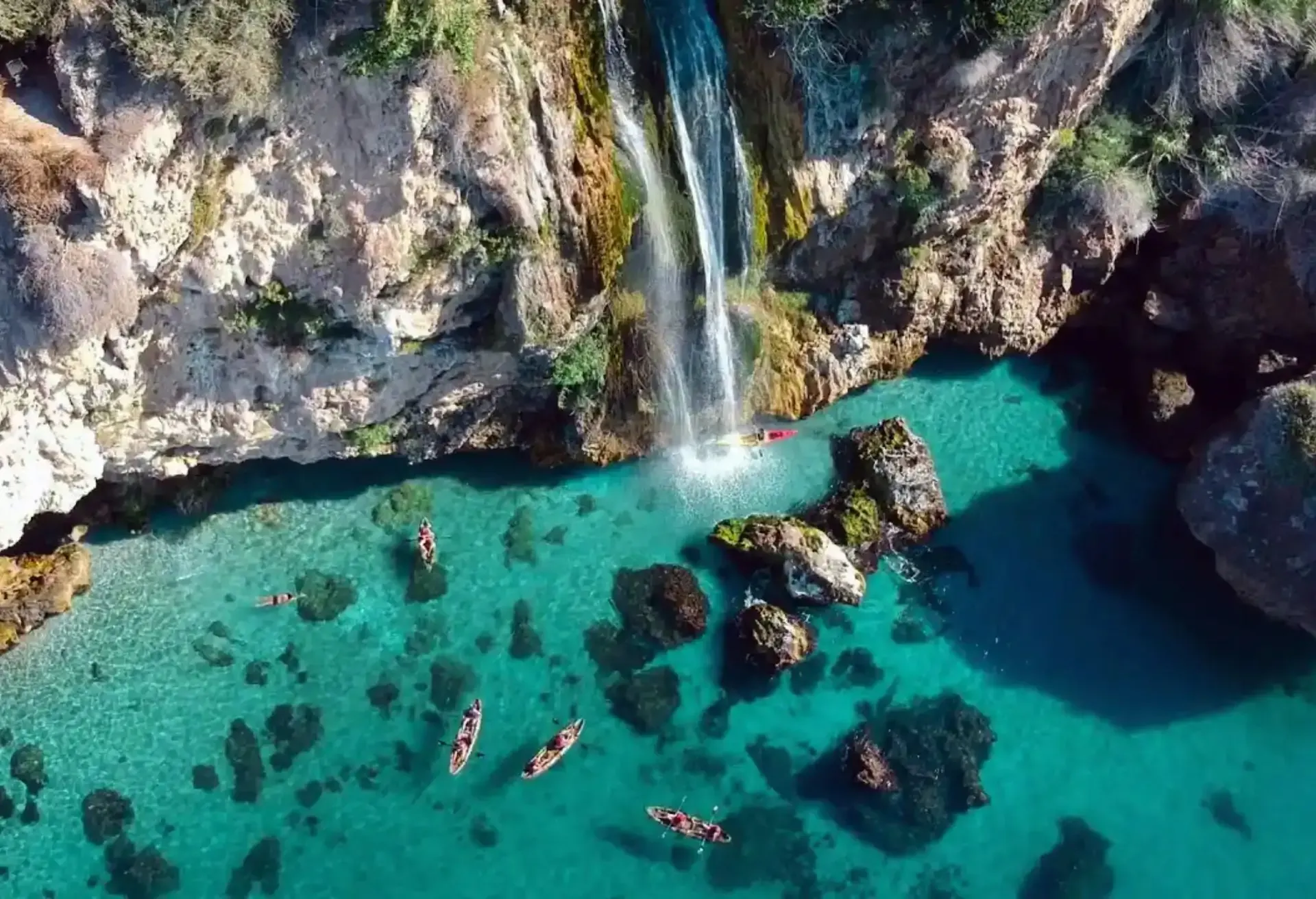 An aerial view of a waterfall cascading from rocky cliffs directly into crystal-clear turquoise waters, with kayakers paddling nearby.