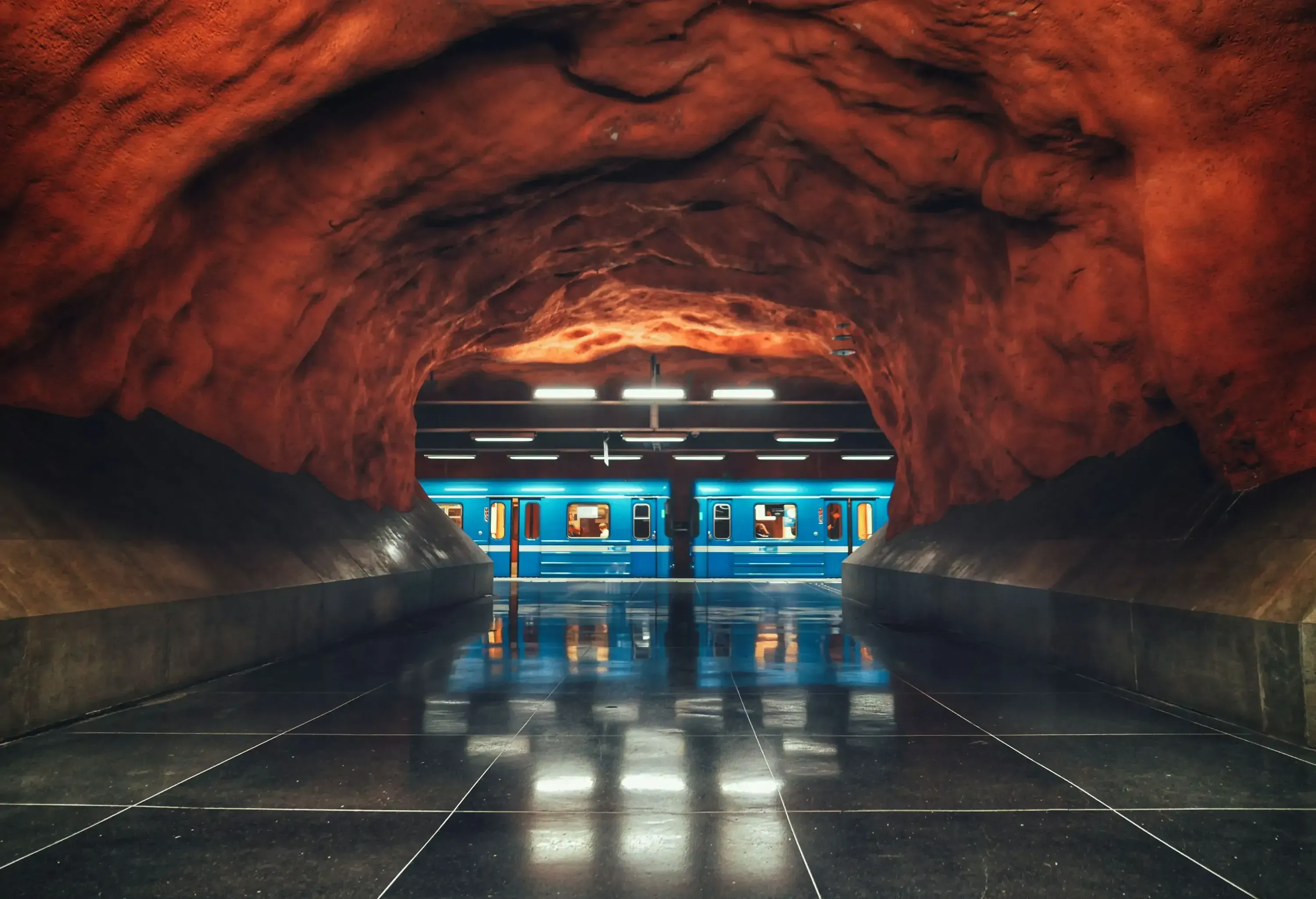 A subway train platform with an arched ceiling of uneven red rocks. 