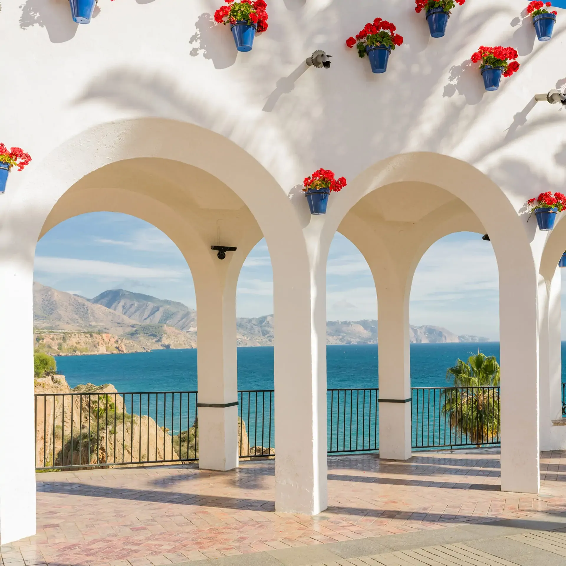 White stone arches decorated with red flower pots on a terrace with views of a turquoise sea and mountainous coastline.