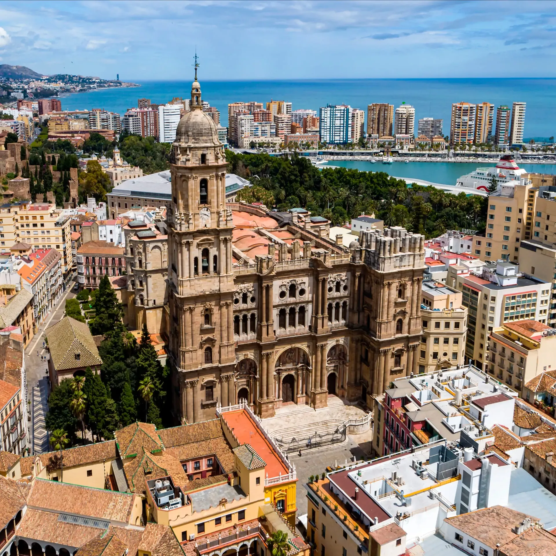 An aerial view of an imposing historic cathedral surrounded by city rooftops, with a harbor and the blue sea in the background.